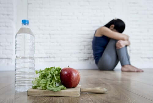 Kvinne som sitter på gulvet og klemmer knærne til brystet med salat, eple og vann i forgrunnen.