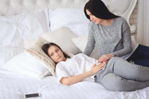 En tenåring som ligger i sengen med hånden på magen, og moren som trøster henne. 