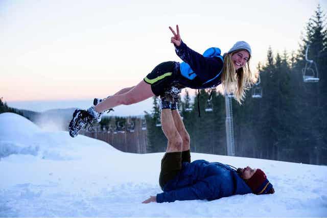 Et blåtonet bilde av en mann og en kvinne som leker i snøen på toppen av en skibakke, med stolheis i bakgrunnen